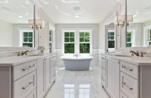 Luxury master bathroom with stand-alone soaker tub, dual vanities with marble counters, brass fixtures, marble floors, and lush nature outside the windows