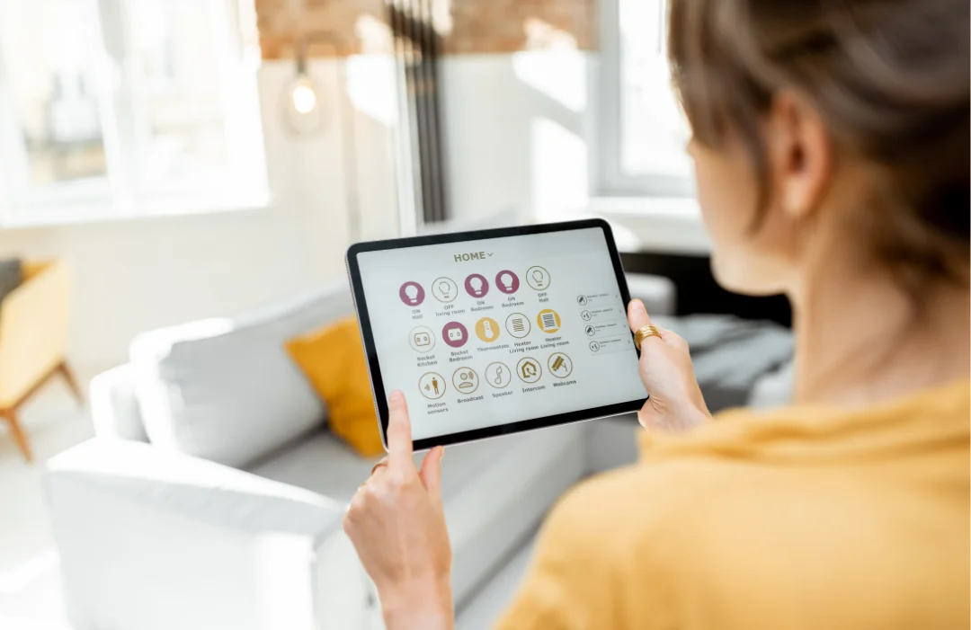 A woman looking at a tablet with smart home icons on the screen while standing in her living room.